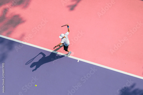 aerial view of a young tennis player in action on a vibrant pink and purple hard court. Dappled tree shadows and strong sunlight create a dynamic and aesthetic summer sports composition.