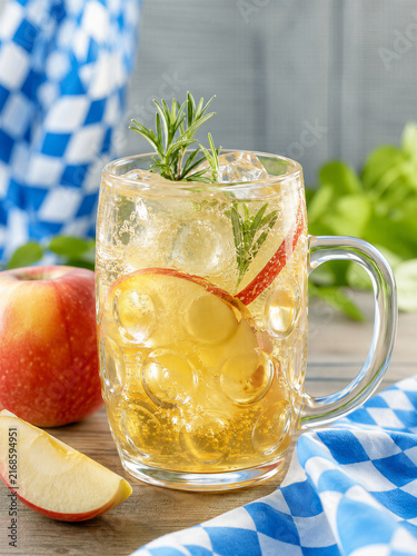 Cold apple cider with ice and rosemary in glass beer mug on wooden table with Bavarian flag. Refreshing autumn beverage for Oktoberfest celebration. Traditional German festival drink concept.