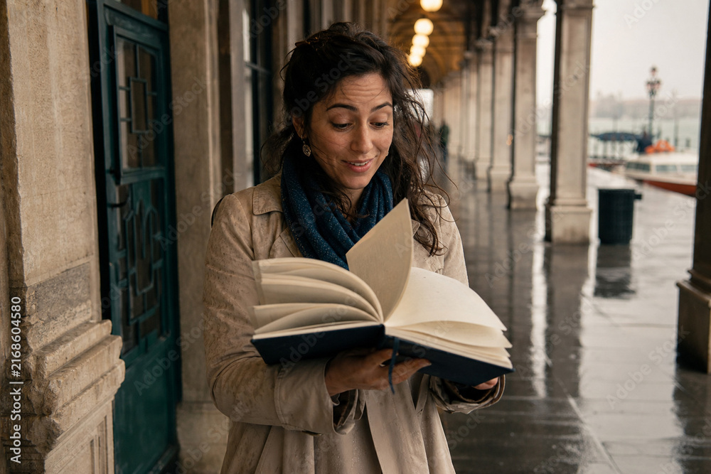 Obraz premium A woman in a beige coat and scarf reads a book along a rainy outdoor colonnade, eyes focused, a gentle smile as pages turn, candid lifestyle moment under cloudy skies.
