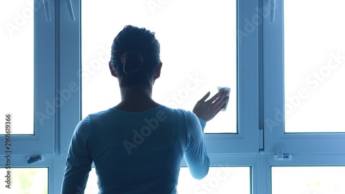A person stands near a window and cleans it with a cloth. The setting is bright, indicating it is daytime. There are several windows in the view.