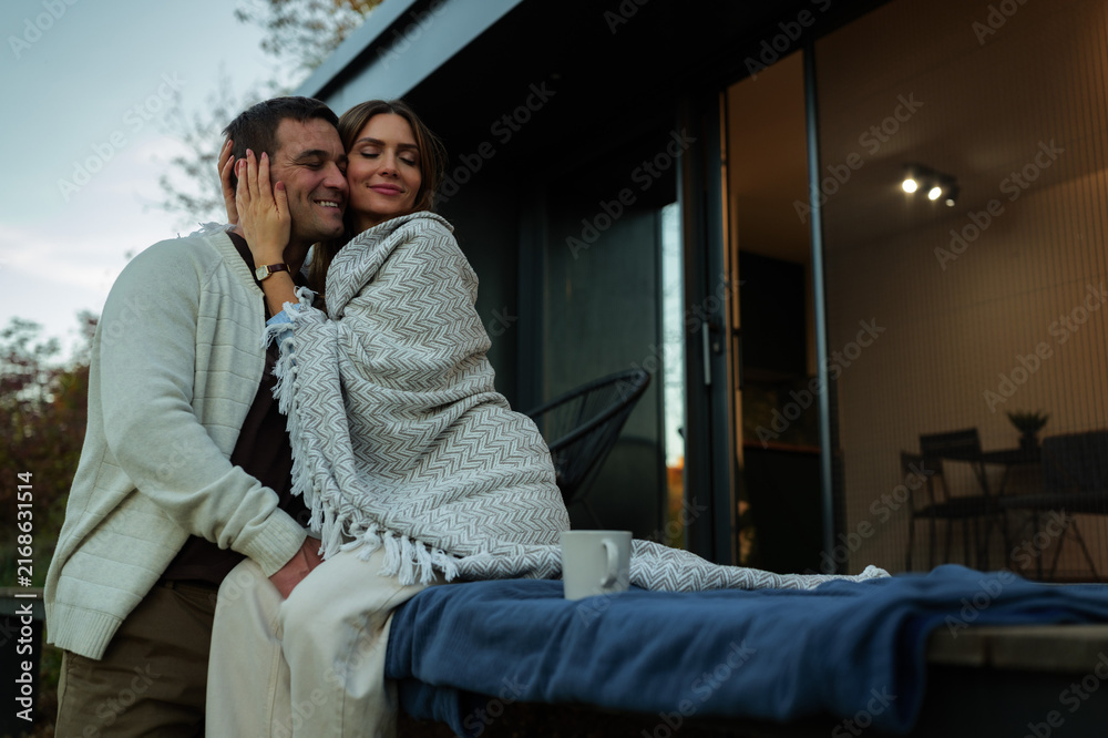 Fototapeta premium Loving couple enjoying a peaceful outdoor moment together, embracing on the patio of their modern home while wrapped in a warm blanket, finding comfort and connection
