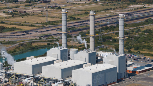 Aerial view of Grain CHP Power Station with its three prominent chimneys, industrial buildings, and steam vents set against the surrounding landscape in Isle of Grain, England, United Kingdom.