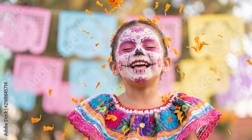 Mexican girl with Catrina-inspired sugar skull makeup and ruffled dress laughing among falling marigold petals, ideal for Day of the Dead promotional visuals and posters.