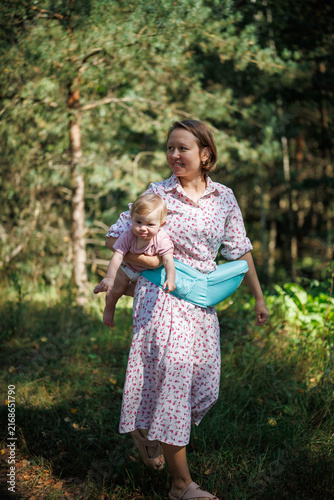Woman carrying a baby in a hipseat while walking through a green forest