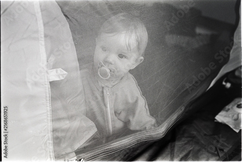 Candid shot on analog black and white film showing a baby with a pacifier looking through a mesh tent door. Visible film grain