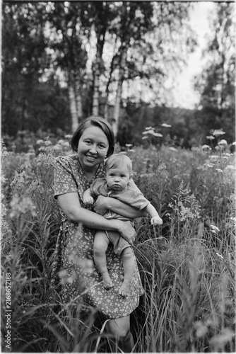 Grayscale film photograph showing a smiling woman holding a baby while standing among tall grasses in a natural setting