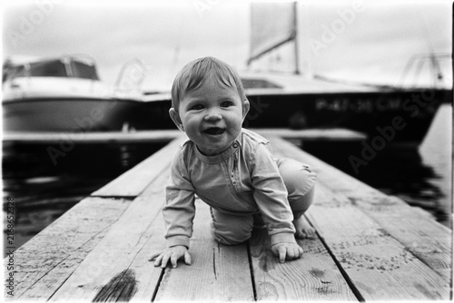 Wide-angle film photograph in black and white of a baby crawling on a wooden pier with boats in the background