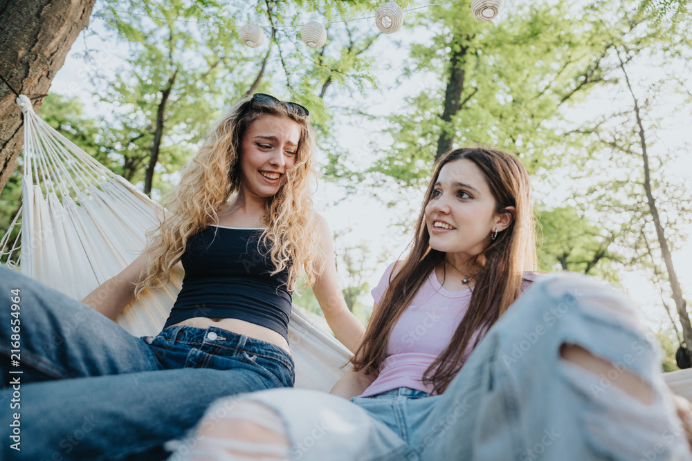 Fototapeta premium Two teenage friends relax and laugh together in a hammock under leafy trees. The casual outdoor scene shows a carefree summer moment between close friends.