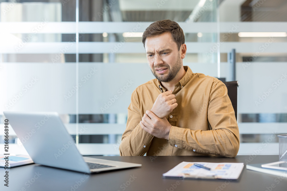 Obraz premium Businessman experiencing acute wrist pain and discomfort while sitting at an office desk and working on a laptop, indicating potential carpal tunnel syndrome or a repetitive strain injury