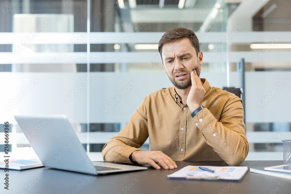Obraz premium Man experiencing dental pain and discomfort while sitting at his office desk, holding his cheek and trying to focus on his laptop during a busy workday