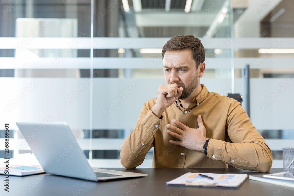 Obraz premium Man feeling unwell and coughing into fist, experiencing illness symptoms while working at his desk in a modern office, highlighting workplace health and the spread of respiratory viruses