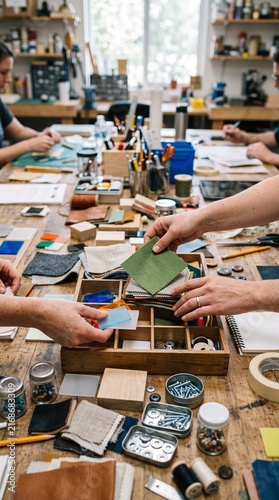 Crafting Together: Hands of artisans meticulously select materials at a cluttered workshop table, demonstrating a collaborative creation process with focus and skill.