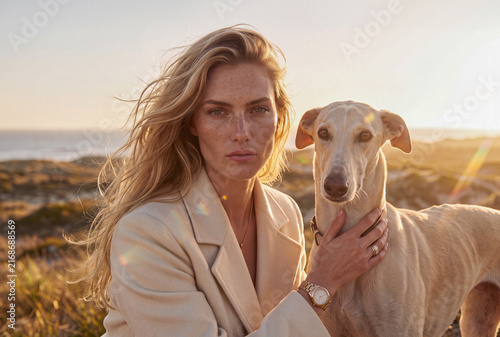 High fashion editorial portrait of a blonde woman with freckles hugging greyhound dog at beach sunset