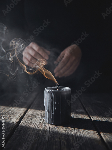 Hand lighting black candle with rising smoke on rustic wooden table, moody ritual still life