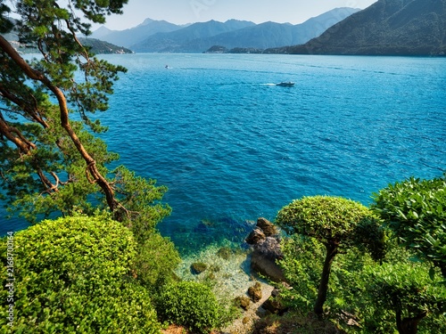 Canvas Print Scenic view of turquoise Lake Como from Villa del Balbianello gardens in Italy, with lush greenery, pine trees, boats and Alpine mountains