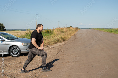 male driver stretching his legs beside a parked car on an empty country road, taking an active break to reduce stiffness and fatigue after prolonged sitting on a long journey