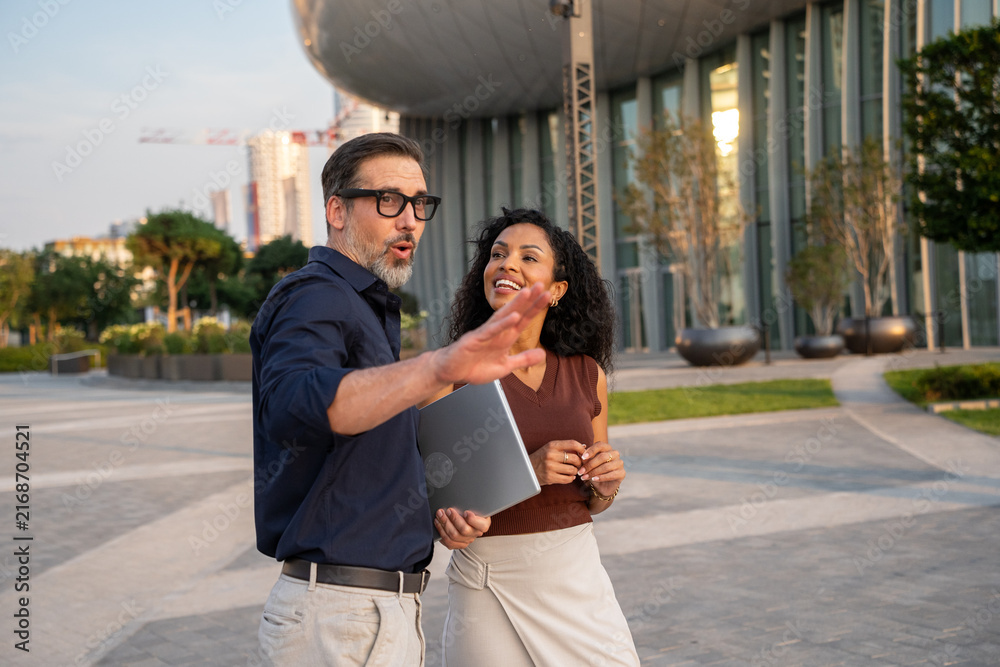 Fototapeta premium Two diverse business professionals standing outside a modern glass office building, looking at a discussing a project during a meeting. 