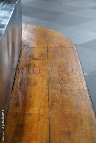 A close-up detail of the smooth teak wood surface of a curved bench, set against a background of symmetrically patterned flooring in a public space.