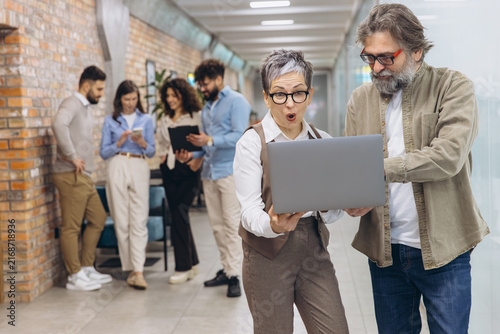 Surprised senior businesswoman and male colleague looking at laptop, reacting to shocking news or amazing results in office hallway