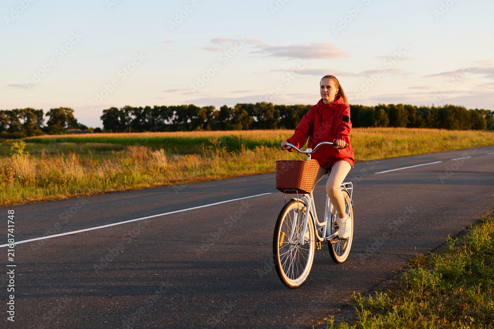 Fototapeta premium A person cycles on a road lined with fields. The sun sets in the background, casting warm light on the scene. The cyclist enjoys the ride in a casual outfit