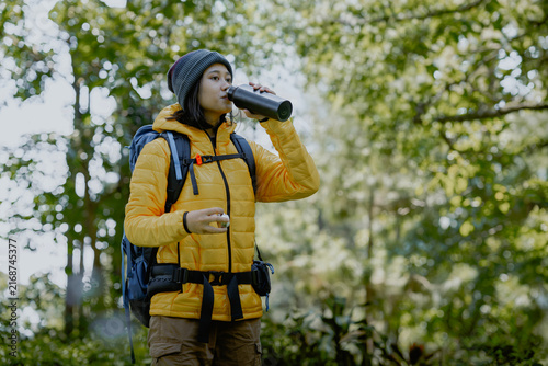 Young asian female hiker drinking water from aluminium bottle in a green forest. Nature, healthy and adventure lifestyle.