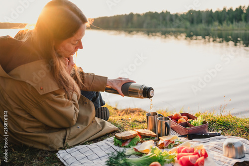 Woman pouring a hot drink from a thermos during an outdoor picnic by a peaceful lake, enjoying fresh food, warm sunset light and relaxing time in nature