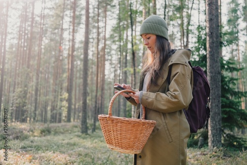 Woman using a smartphone while holding a wicker basket in a pine forest during a seasonal foraging trip, combining modern technology with mushroom hunting
