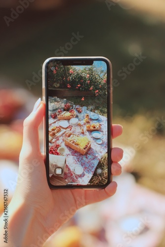 concept of sharing moments on social media. Gen Z lifestyle. Person holding smartphone capturing photo of picnic basket filled with fresh fruits and vegetables outdoors