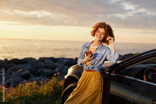 Cheerful woman with curly hair leaning on a car by the sea at sunset, glancing at her phone. Casual lifestyle shot emphasizing outdoor freedom, travel, and relaxed moments.