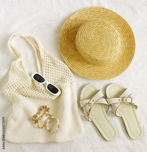 Flat lay of women's casual summer outfit neutral colors. Top view straw hat, bag, sunglasses, shoes, bracelets on white linen background. Lifestyle .Space for text.