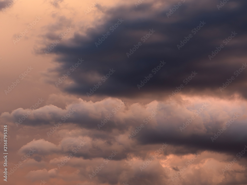 Fototapeta premium Dramatic dark storm clouds illuminated by warm orange sunset light, creating a moody atmospheric cloudscape before a possible thunderstorm.
