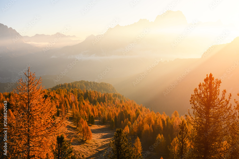 Naklejka premium Sun rays stream through mountain haze above golden larch forests in the Dolomites. Atmospheric autumn landscape with warm backlight and layered alpine ridges