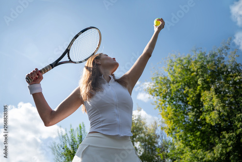 Young Caucasian female tennis player raising ball and racket to serve against blue summer sky. Athletic performance, competition, strength and active outdoor sport concept.