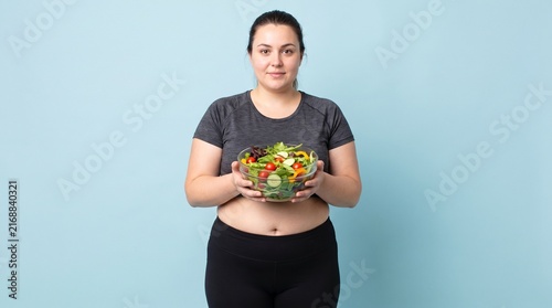 An overweight adult woman with visible abdominal fat standing upright while holding a bowl of fresh vegetables, representing dieting, healthy nutrition, and weight loss, isolated against a plain pale 