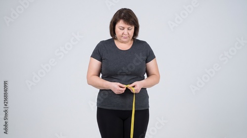 An overweight adult woman standing front-facing while measuring her waist with a flexible measuring tape, visibly highlighting abdominal fat and weight management, wearing simple sportswear, isolated 