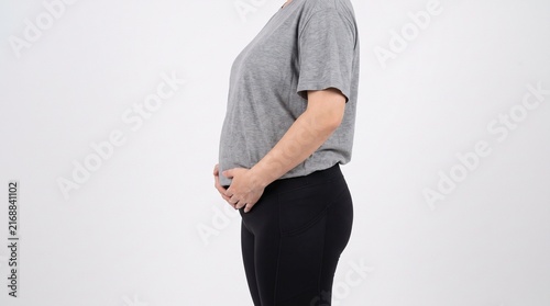 An overweight adult woman shown in side profile gently holding her abdominal area to highlight visible belly fat and excess weight, wearing simple neutral athletic clothing, isolated against a plain 