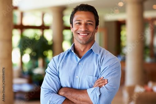 Smiling man in a blue button down shirt standing with arms crossed in a bright hotel lobby