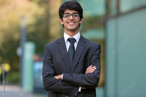 Young man in a dark suit and tie standing with arms crossed outdoors in a city setting
