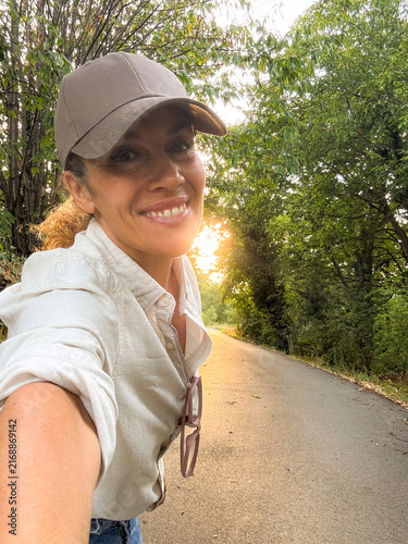 Smiling woman taking a sunset selfie on a quiet country road, casual cap and shirt, sunglasses hanging, enjoying nature and a peaceful evening walk