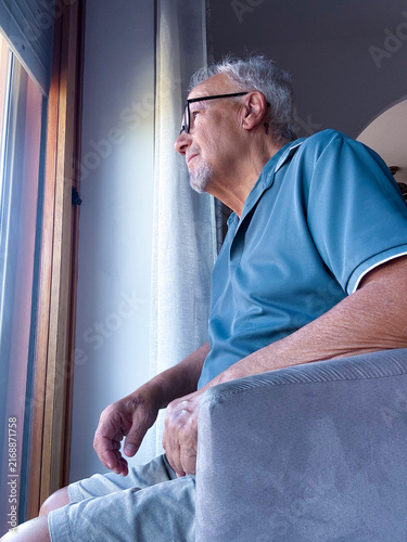 Bild auf Leinwand Elderly man sitting in a chair looking thoughtfully out a window at home, contem