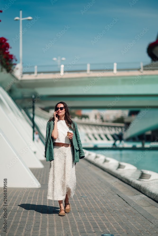 Fototapeta premium Woman walking, talking on smartphone, holding coffee cup in sunny valencia, spain, modern city street background