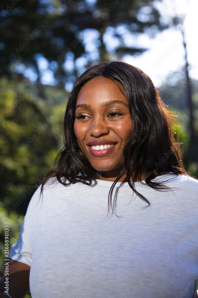Fototapeta premium African adult woman smiling while standing in woodland wearing white t-shirt, showing sunlit bokeh