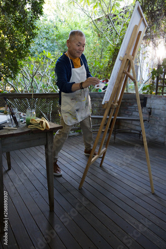 Foto Senior Asian man painting on wooden deck wearing paint-stained apron with easel