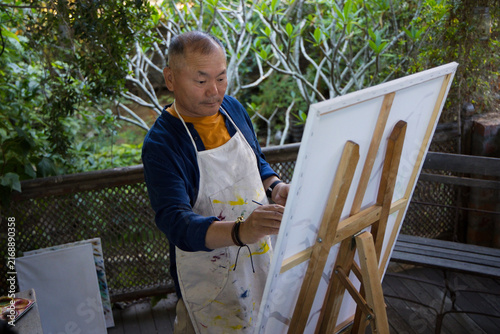 Obraz na plátne Senior Asian man standing and painting at wooden easel on garden deck wearing pa