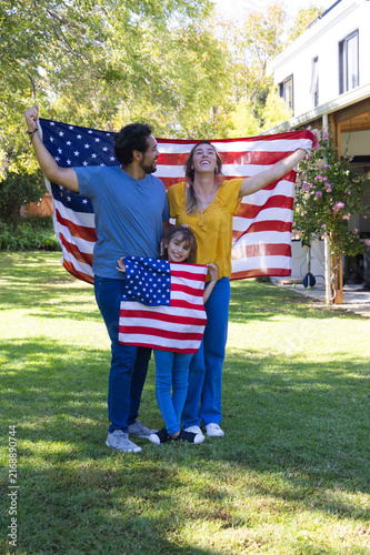 Diverse family standing on suburban lawn holding large American flag, youth holding small flag