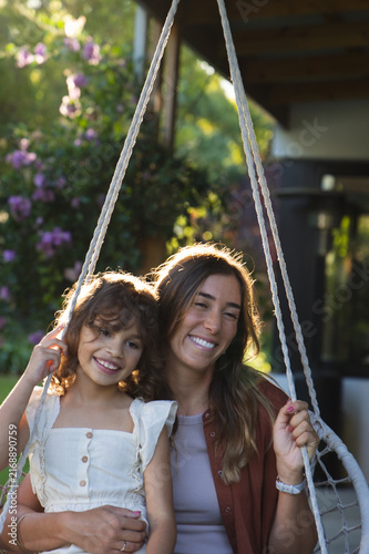 Diverse mother and daughter sitting on hanging rope swing chair at porch, holding braided ropes