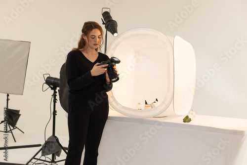 Woman in black outfit holding DSLR camera and reviewing shots of skincare bottles in studio softbox