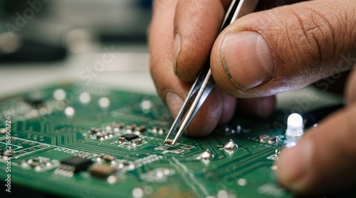 Close up of a technician's hands using tweezers to place a small component onto a green circuit board
