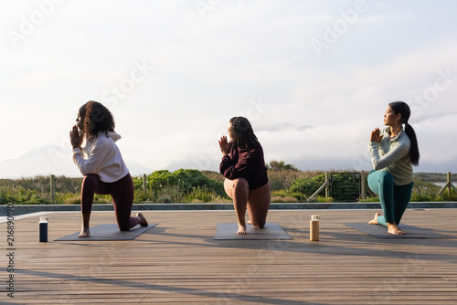African women practicing kneeling lunge in hoodies, leggings on coastal deck with mats, bottles