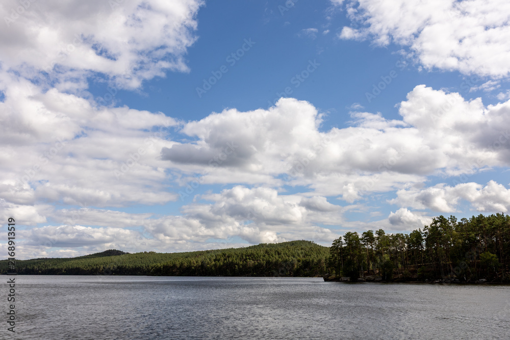 Fototapeta premium Scenic lake landscape with calm rippling water, lush green shoreline, resort buildings and gazebos surrounded by trees, with mountains visible in the distance under a dramatic cloudy sky. Peaceful out